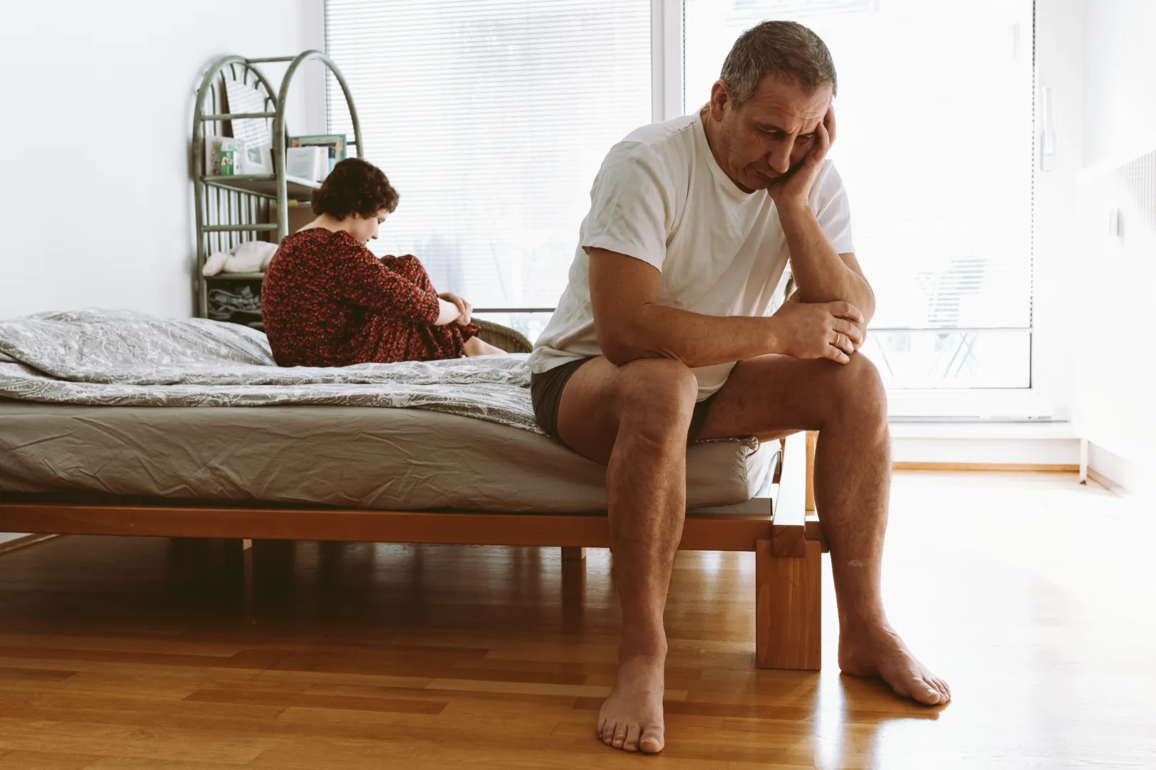 Man sitting on the edge of a bed with his head in his hand, woman sitting behind him — symbolic of concerns around erectile dysfunction and medication side effects.